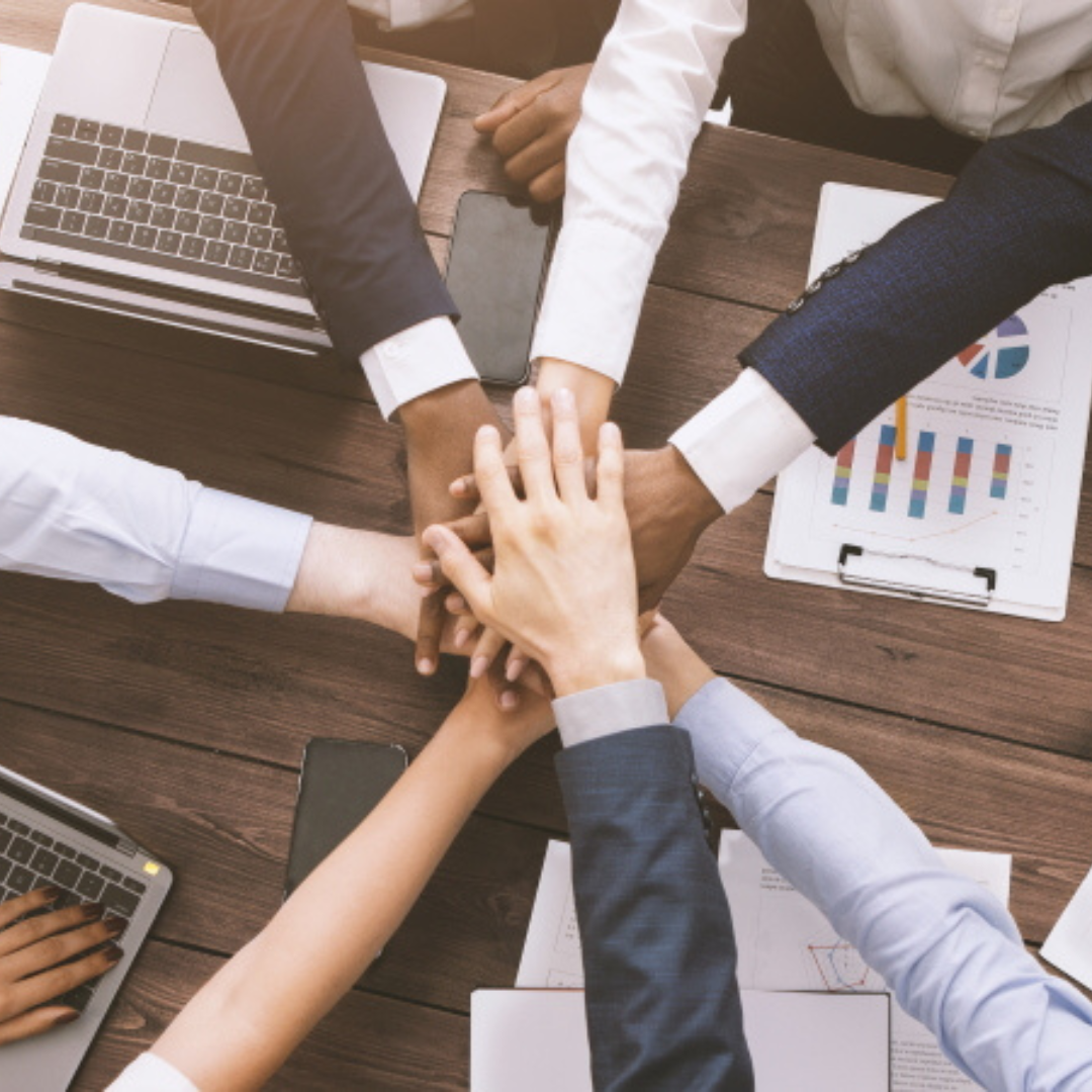 A diverse group of business professionals in formal attire, putting their hands together in a show of unity and teamwork. Laptops, charts, and a phone are visible on the wooden table.</p>
<p>