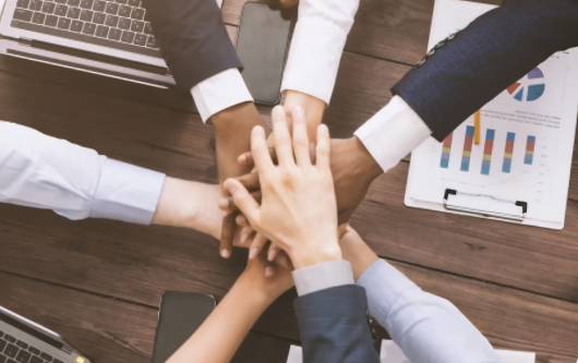 A diverse group of business professionals in formal attire, putting their hands together in a show of unity and teamwork. Laptops, charts, and a phone are visible on the wooden table.</p>
<p>